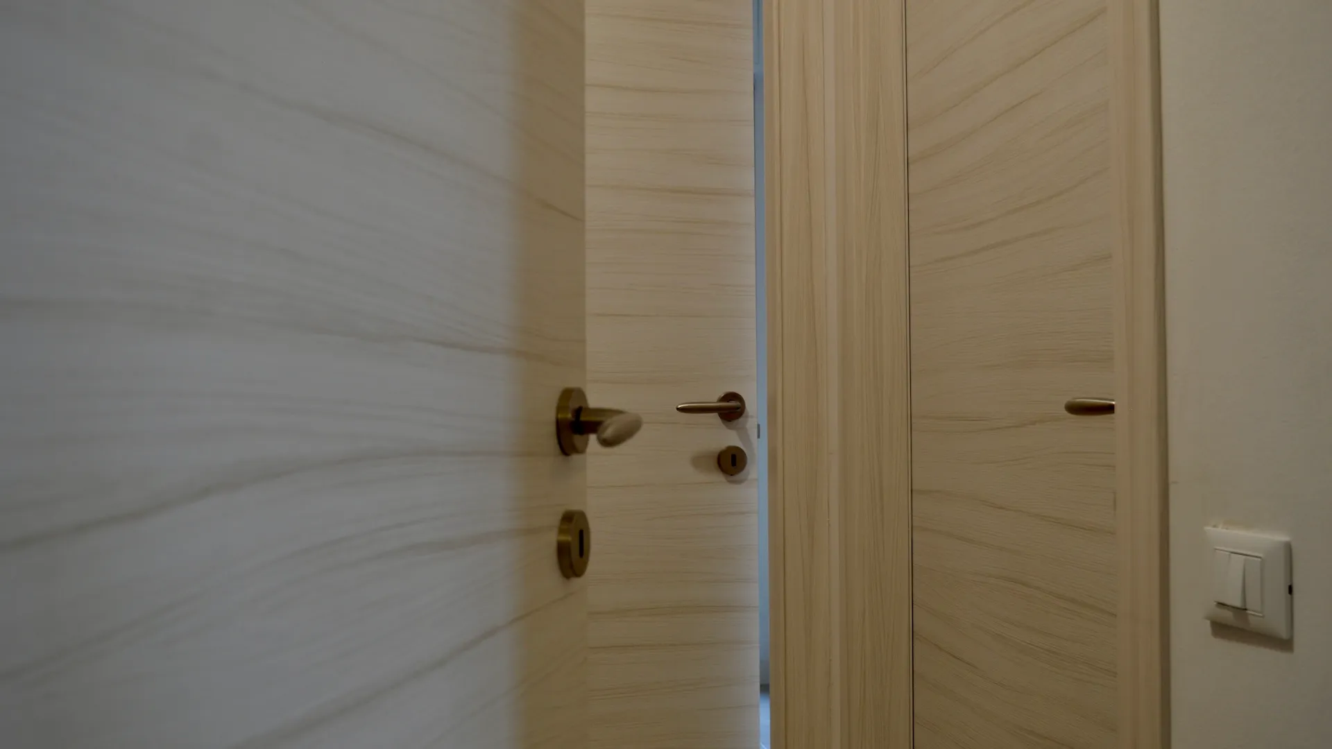 Porta interna in legno chiaro socchiusa su un corridoio, con maniglia e serratura in ottone